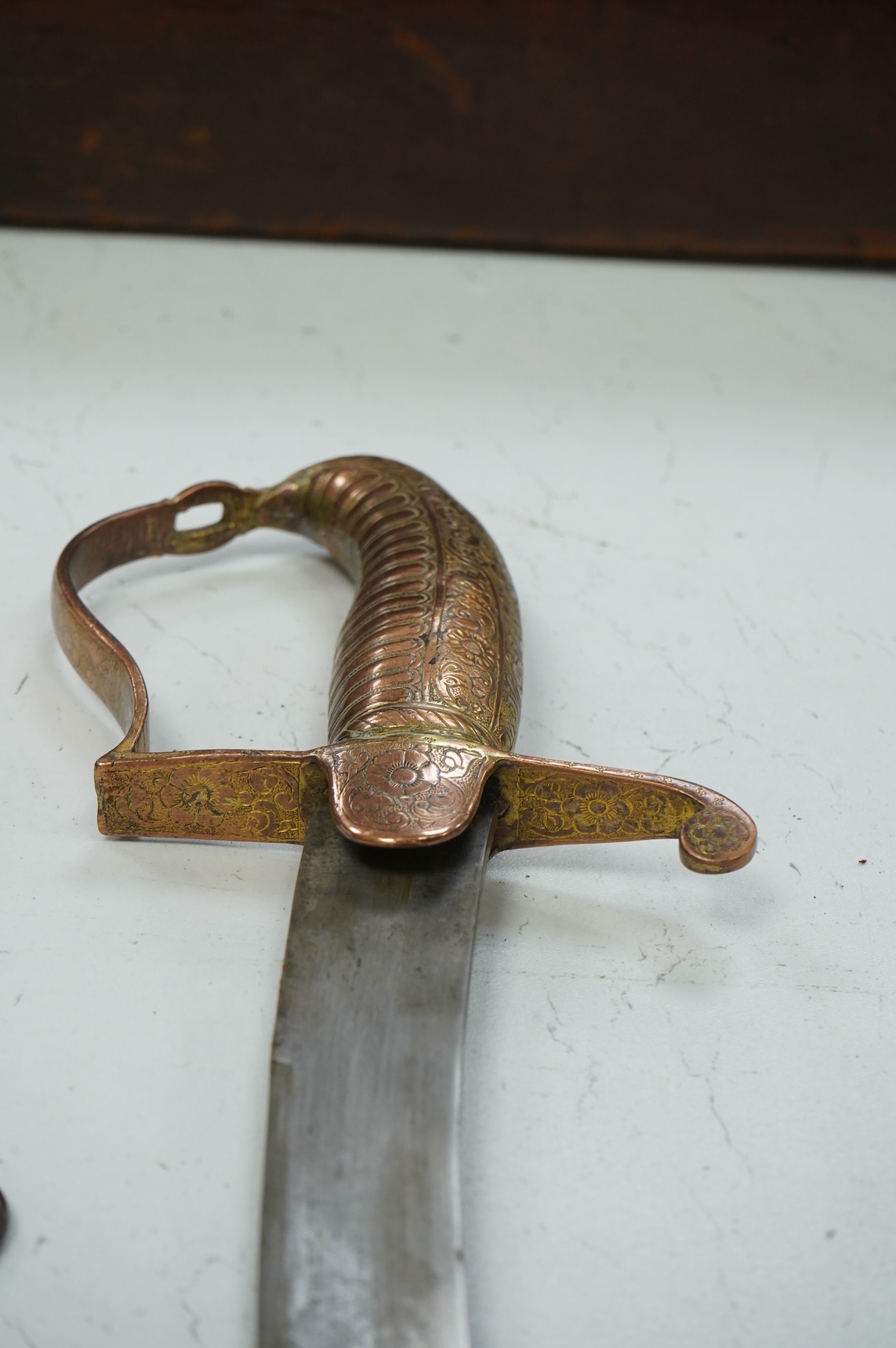 An early 19th century Indian sabre with curved single edge blade, copper grip and stirrup hilt, in its copper scabbard embossed with lions, scrolling foliage and flowers